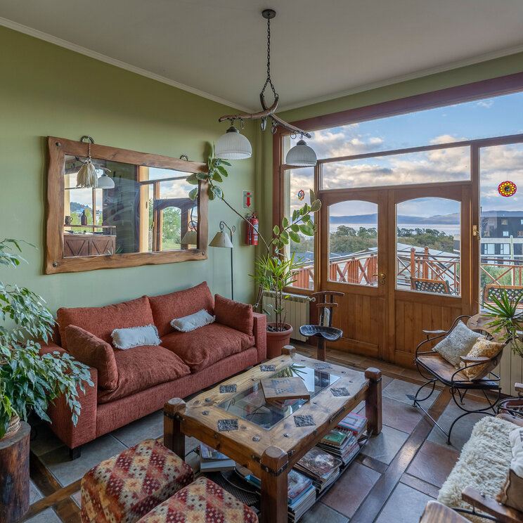 Gemütlicher Wohnbereich im Hotel Tierra de Leyendas in Ushuaia mit rotem Sofa, rustikalem Holztisch und Panoramafenster.