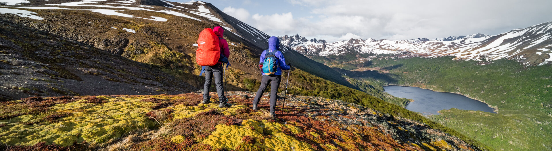 Zwei Wanderer mit Rucksäcken blicken auf einen See und schneebedeckte Berge in Patagonien.