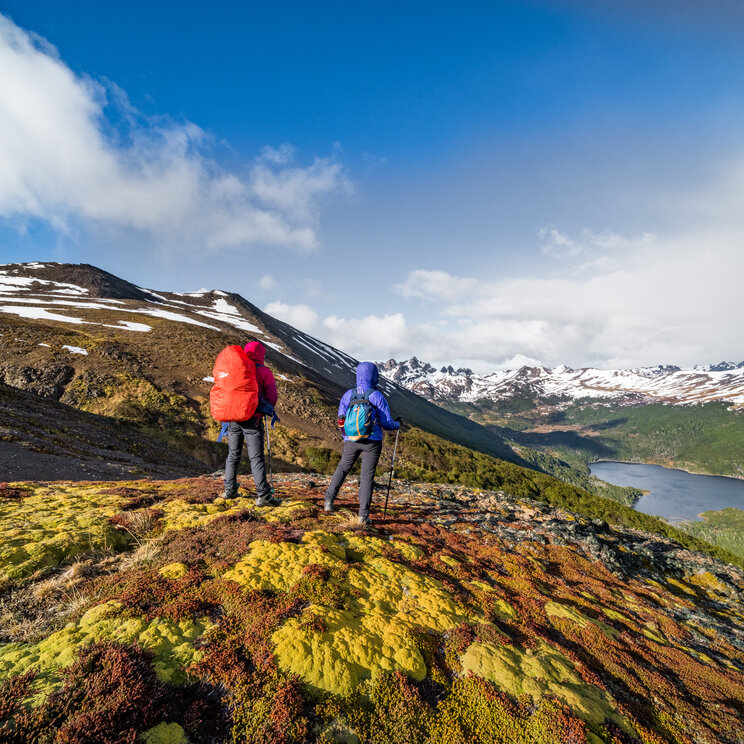 Zwei Wanderer mit Rucksäcken blicken auf einen See und schneebedeckte Berge in Patagonien.