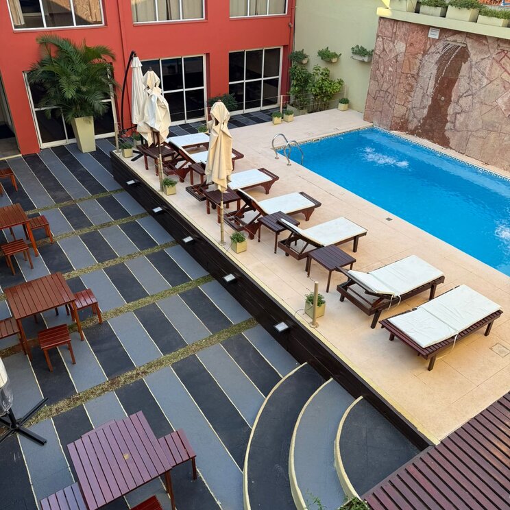 Aussenpool mit Sonnenliegen, Schirmen und Sitzbereichen mit Holztischen im Innenhof eines roten Hotelgebaeudes in Iguazo