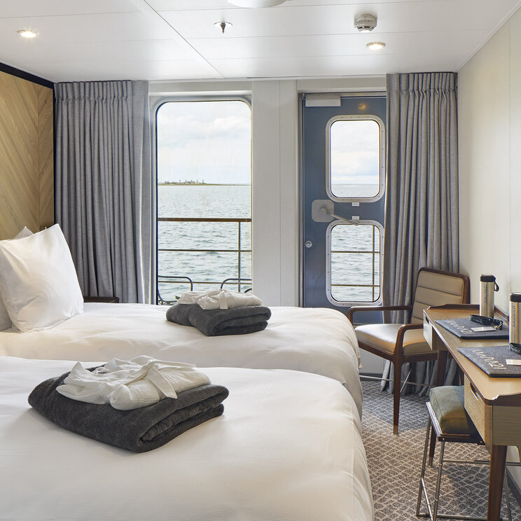 Elegante, modern eingerichtete Balkonkabine an Bord des Expeditionsschiffs Magellan Explorer mit Doppelbett, hellen Holzmöbeln und direktem Blick durch das Panoramafenster auf die antarktische Eislandschaft.