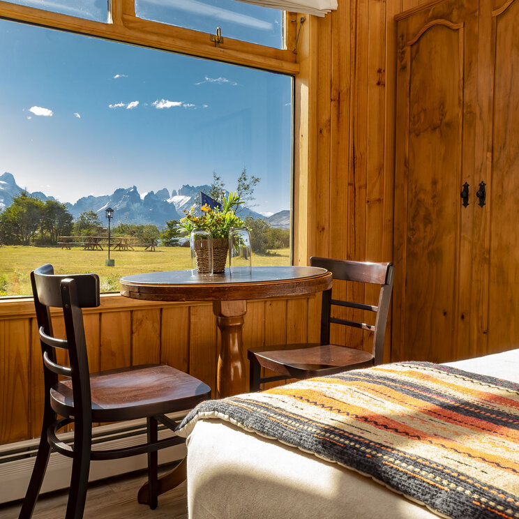 Sitzbereich im Doppelzimmer mit Holztisch und Blick durch ein Panoramafenster auf das Torres-del-Paine-Massiv.