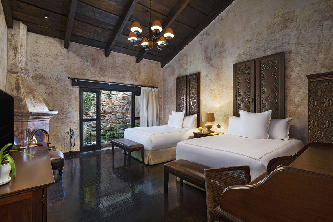Stilvolles Zimmer im El Convento Boutique Hotel in Antigua, Guatemala, mit zwei Betten, Kamin, edlem Holzdekor und Blick in den Innenhof – luxuriöse Unterkunft im kolonialen Stil nahe der Altstadt.