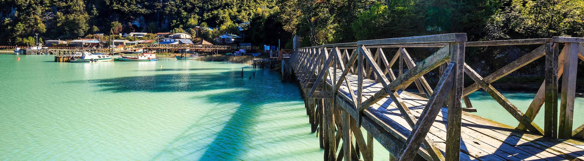 Holzsteg über türkisblauem Wasser in Caleta Tortel. Im Hintergrund sieht man Jachten und Wald
