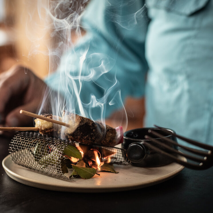 Restaurant im Patagonia Camp in Torres del Paine mit angerichtetem Gourmetgericht auf einem Teller, das am Tisch mit Rauch und offener Flamme präsentiert wird.