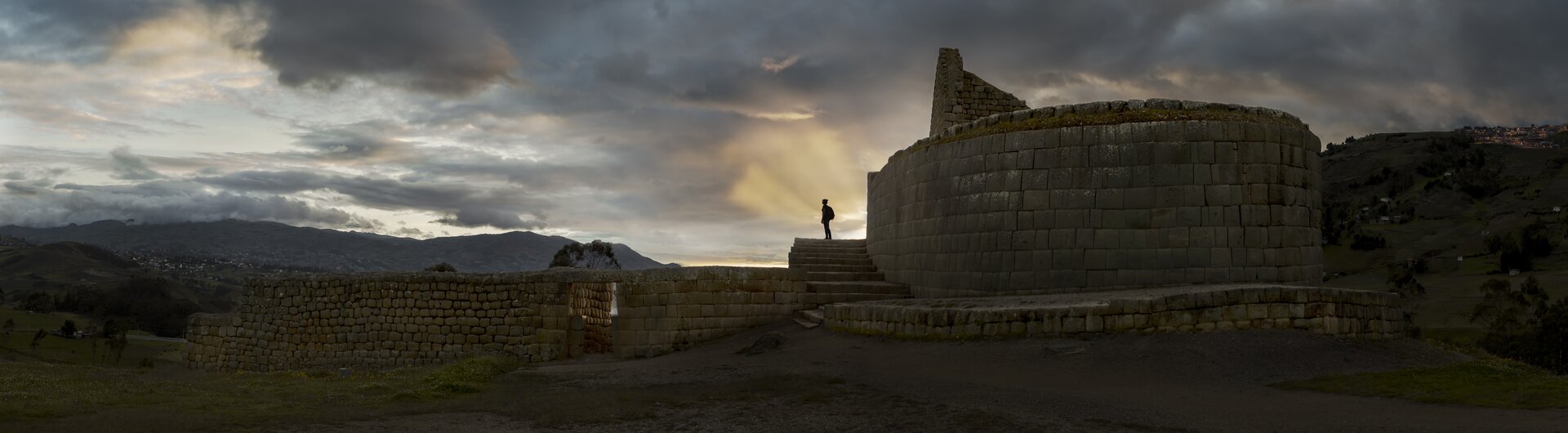 Die runden Inka-Steinmauern des Sonnentempels von Ingapirca in Ecuador, auf denen eine Silhouette vor dramatischem Wolkenhimmel im Sonnenuntergang steht.