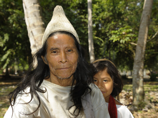 Porträt einer Person mit spitzem Hut und einer weiteren Person im Hintergrund vor Bäumen in Tayrona, Kolumbien.