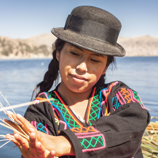 Eine Frau mit schwarzem Hut flicht Totora-Schilf auf einer Insel am Titicacasee in Peru vor blauem Wasser.