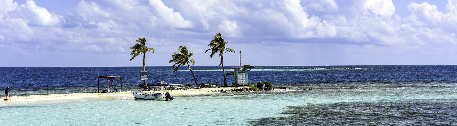 Kleine Sandinsel Silk Caye mit drei Palmen, Boot und Holzsteg im türkisfarbenen Wasser des Karibischen Meers.