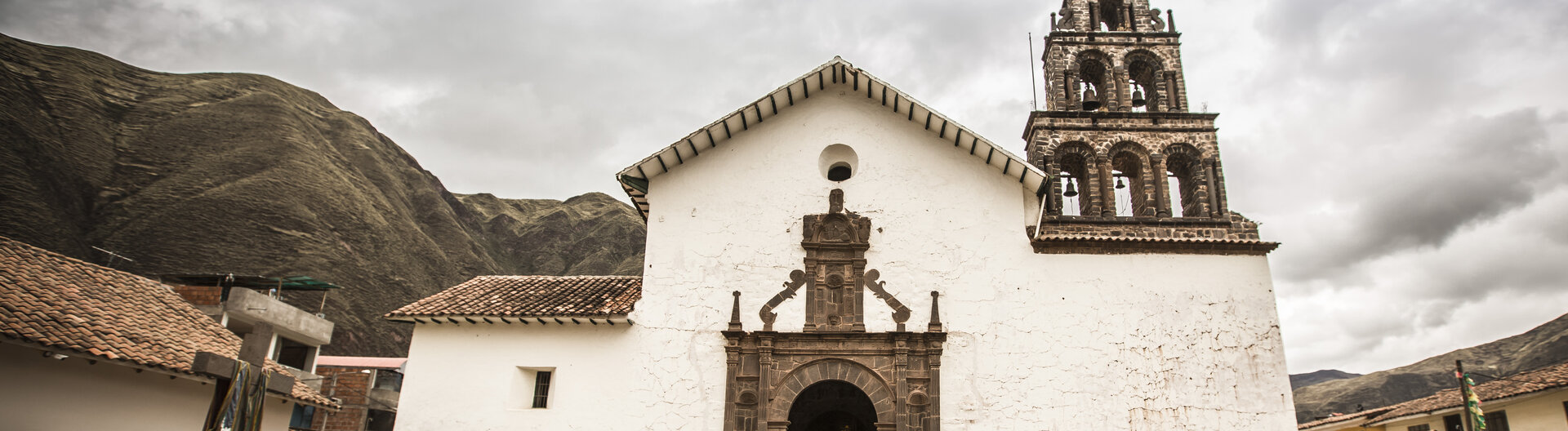 Die weiße Kolonialkirche San Juan Bautista in Huaro mit Steinturm vor Bergen unter bewölktem Himmel in Peru.