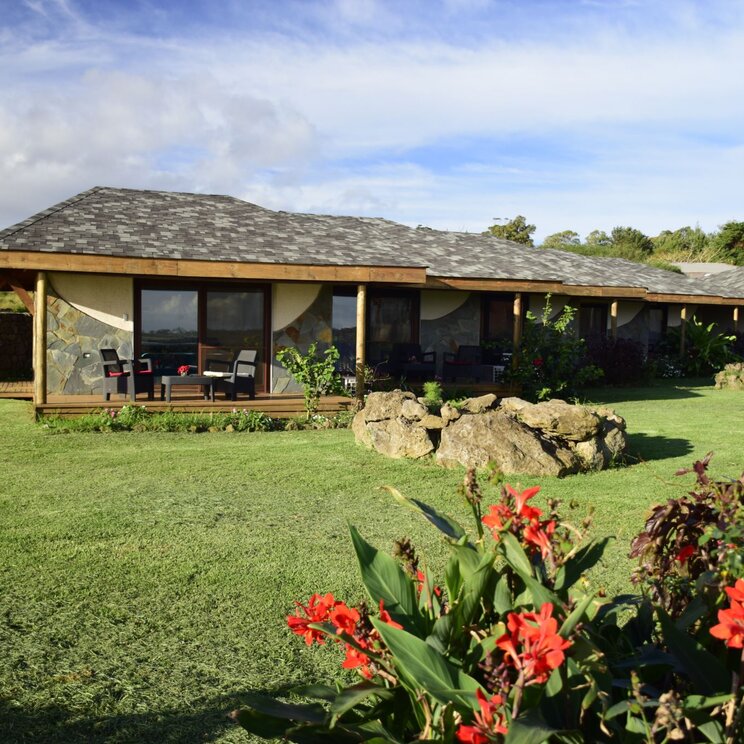 Außenansicht der Takarua Lodge mit Steinfassade, Rasenfläche und roten Blumen im Vordergrund, Osterinsel.