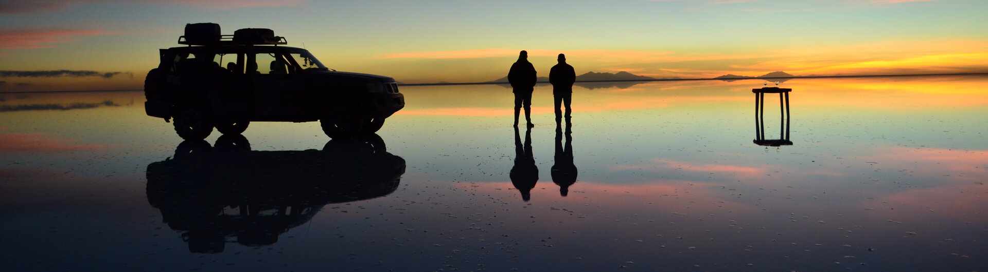 Silhouetten von zwei Personen, Geländewagen und Tisch spiegeln sich im gefluteten Salar de Uyuni in Bolivien bei Sonnenuntergang.