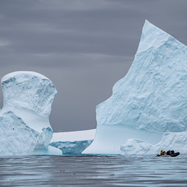 Ein kleines Schlauchboot mit Passagieren fährt im Wasser an riesigen, steilen Eisbergen unter grauem Himmel in der Antarktis.