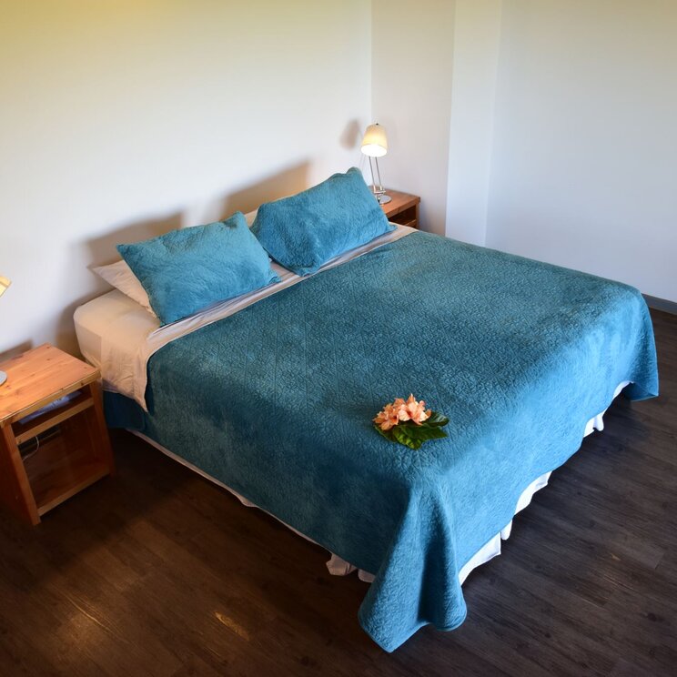 Doppelzimmer mit dunklem Holzboden, türkisblauem Bettüberwurf und einer Blume als Dekoration, Osterinsel.