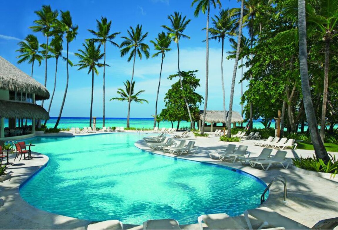 Geschwungener Außenpool mit hellblauem Wasser, weißen Liegen und hohen Palmen vor dem Meer in Punta Cana.