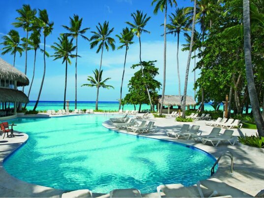 Geschwungener Außenpool mit hellblauem Wasser, weißen Liegen und hohen Palmen vor dem Meer in Punta Cana.