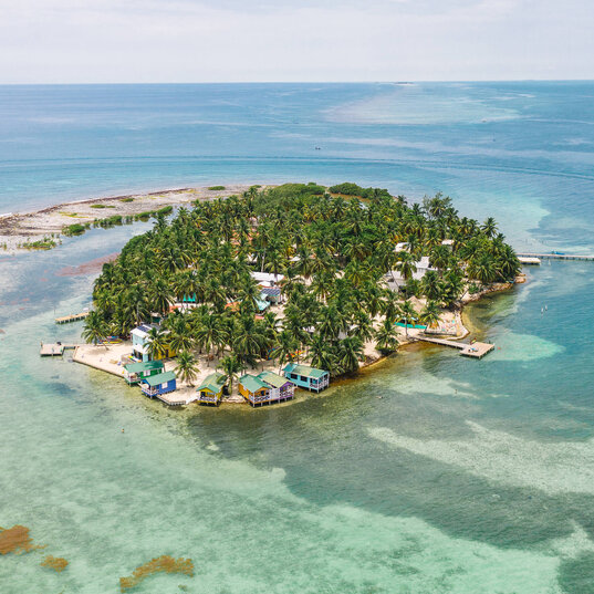 Luftaufnahme der kleinen Insel Tobacco Caye in Belize mit dichtem Palmenbewuchs, bunten Hütten am Sandstrand und umgeben von türkisfarbenem Wasser mit Korallenriffen