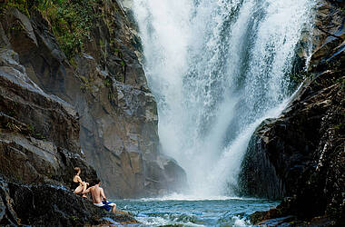 Zwei Menschen sitzen in Badesachen auf den Steinen und bestaunen Big Rock Wasserfall in Belize