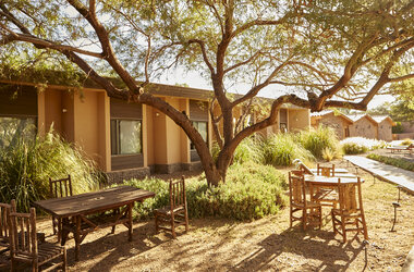 Holztische und Stuehle im schattigen Garten unter Baeumen vor den Hotelgebaeuden in San Pedro de Atacama.