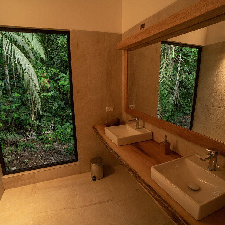 Badezimmer mit Dusche, hellen Fliesen und Blick durch ein Panoramafenster in den Wald, Sacha Lodge.