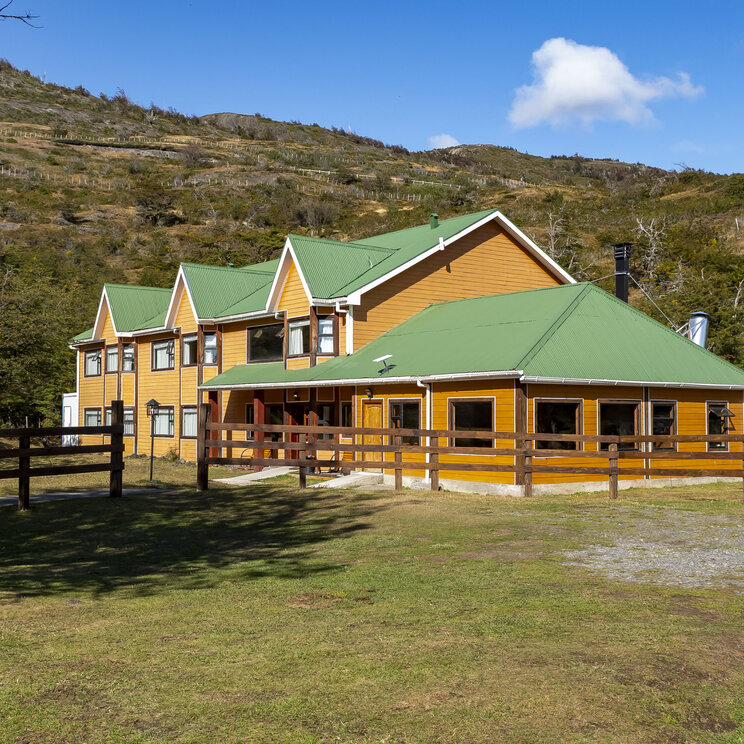 Gelbes Hauptgebäude des Hotel del Paine mit grünen Dächern und Holzzaun vor einer Berglandschaft im Nationalpark Torres del Paine in Patagonien.