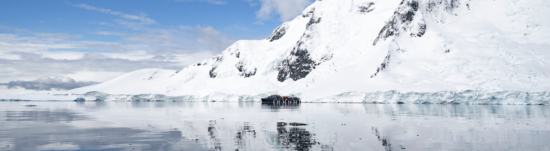Ein Expeditionsschiff ankert vor schneebedeckten Bergen, die sich im ruhigen, blauen Meerwasser der Antarktis spiegeln.