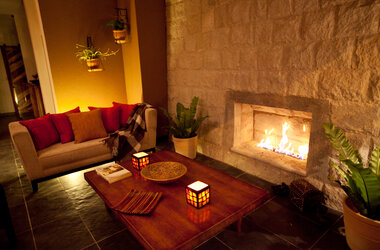 Gemütlicher Loungebereich mit brennendem Kamin in einer Steinwand, Sofa mit roten Kissen und Holztisch.