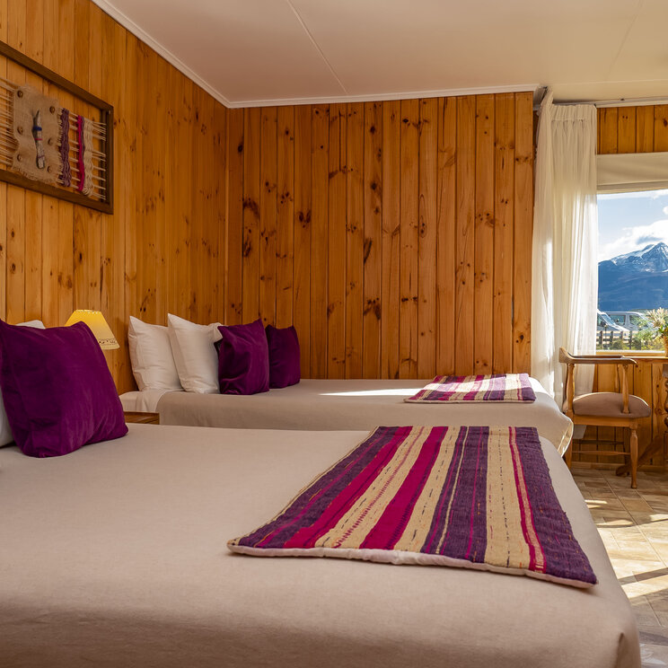 Mehrbettzimmer im Hotel del Paine mit Holzverkleidung, zwei Doppelbetten, bunten Kissen und Fenster mit Bergblick in Chile.