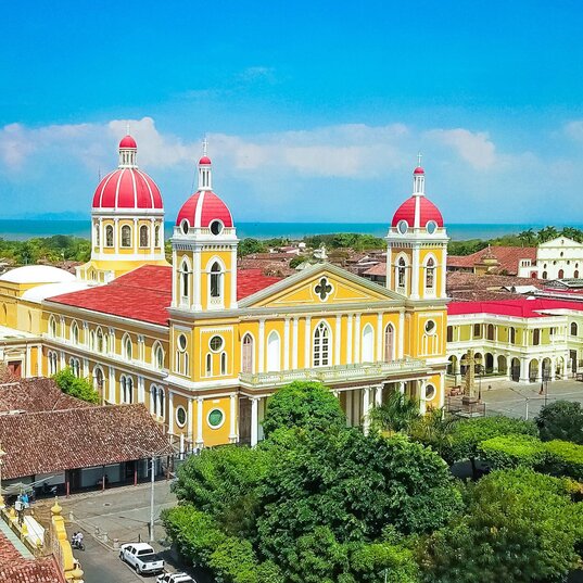 Blick über die Dächer von Granada auf die ikonische gelbe Kathedrale mit ihren roten Kuppeln. Im Hintergrund erstreckt sich der Nicaraguasee.