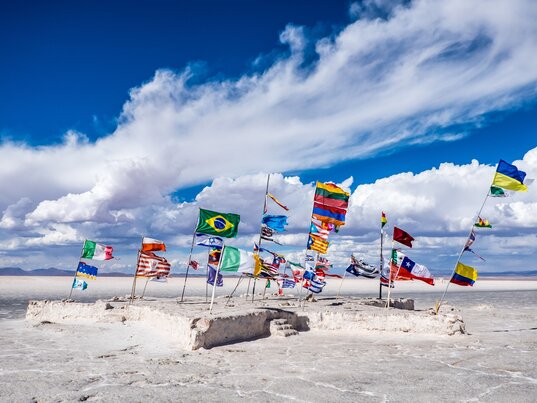 Internationale Flaggen auf einem Salzpodest im Salar de Uyuni in Bolivien unter blauem Himmel mit Wolken.