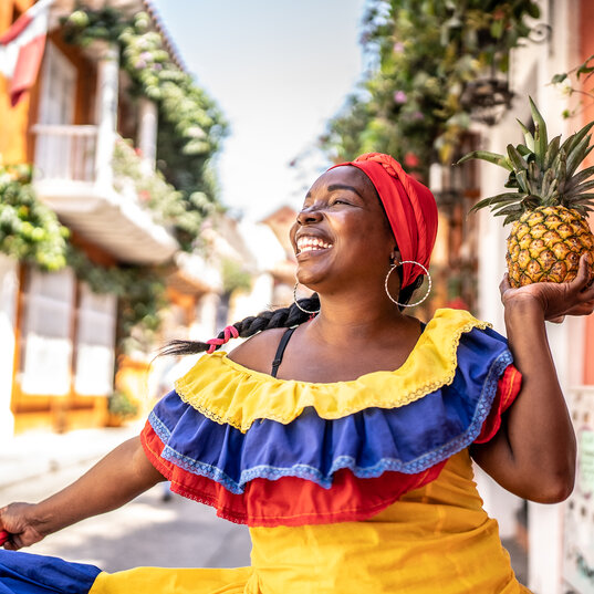 Palenquera in traditioneller gelb-blau-roter Kleidung hält eine Ananas in der Altstadt von Cartagena.