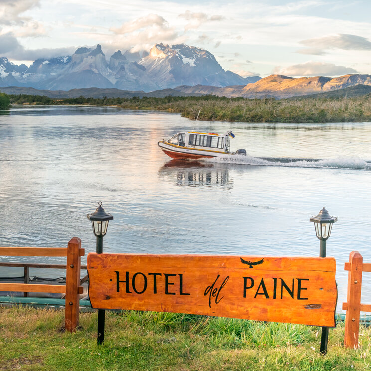 Holzschild des Hotel del Paine an einem Zaun vor einem Fluss mit vorbeifahrendem Boot und Bergen im Hintergrund.