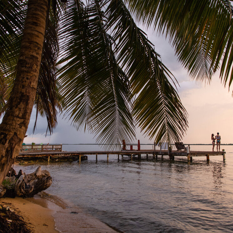 Palmenstrand der Tobacco Caye Paradise Lodge in Belize bei Sonnenuntergang, mit zwei Personen auf einem Holzsteg über dem ruhigen Meer.
