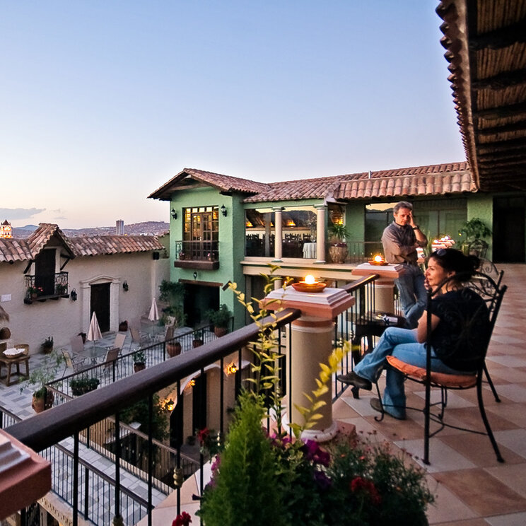 Personen auf einer Terrasse mit Balustrade und Blick auf die Daecherebene von Sucre bei Abenddaemmerung.