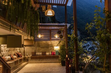 Beleuchtete Außenterrasse mit Sitzbänken, bunten Kissen und Laternen am Flussufer in Aguas Calientes.