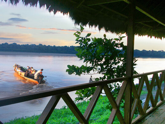 Blick von einer Holzterrasse auf motorisierte Holzboote auf einem breiten Fluss bei Sonnenuntergang in Peru.