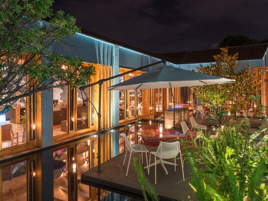Abendliche Terrasse im Hotel Bo mit Sonnenschirm, Sitzplatz auf einem Podest im Wasser und beleuchteten Bäumen.