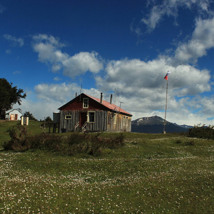 Estancia Kanasaka auf der Isla Navarino in Patagonien, Chile, mit rustikalem Holzhaus, wehender chilenischer Flagge und Blick auf schneebedeckte Berge unter blauem Himmel.