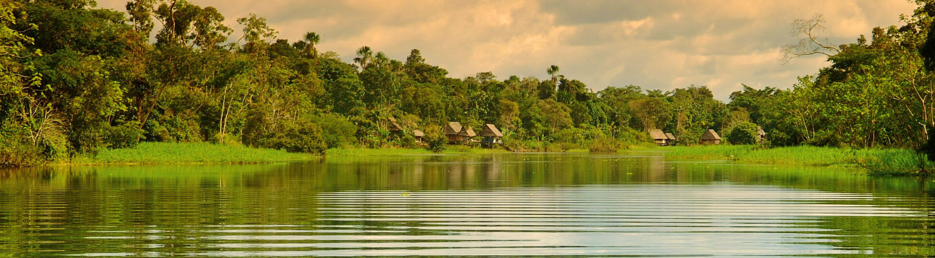 Ein ruhiger Flusslauf im peruanischen Amazonasgebiet, gesäumt von dichtem Regenwald und Hütten unter einem wolkenverhangenen, warmen Himmel.