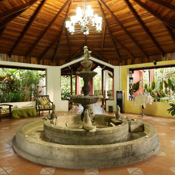 Offene Lobby im Hotel Arenal Paraíso Resort & Spa in La Fortuna, Costa Rica, mit Brunnen, Sitzgelegenheiten und tropischer Bepflanzung unter einem Holzdach.