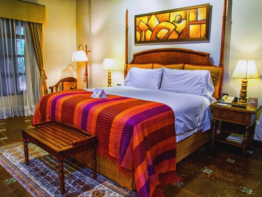 Hotelzimmer in Antigua mit Holzpfeilerbett, bunter Textildecke, gemustertem Teppich und abstrakter Kunst.