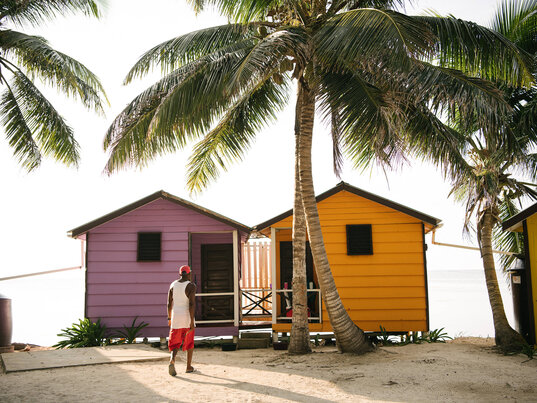 Person mit roter Kappe geht auf zwei bunte Holzhütten am Strand der Tobacco Caye Paradise Lodge in Belize zu, umgeben von hohen Palmen mit Blick auf das Meer.