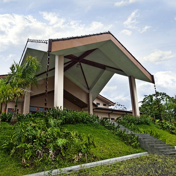 Eingangsbereich des Hotel Arenal Paraíso Resort & Spa in La Fortuna, Costa Rica, mit markanter Dachkonstruktion und umgeben von tropischer Bepflanzung.