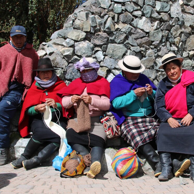 Indigene Frauen in traditioneller Kleidung beim Stricken am Chimborazo in Ecuador, neben einer Steinmauer.