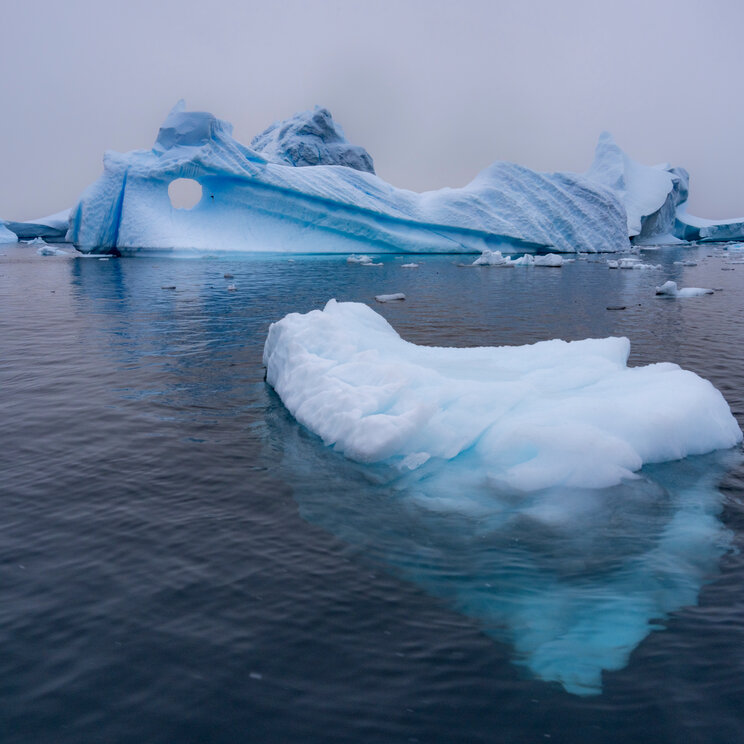Ein kleinerer Eisberg treibt im Vordergrund im Meer vor einer großen Eisformation mit natürlichem Loch in der Antarktis.