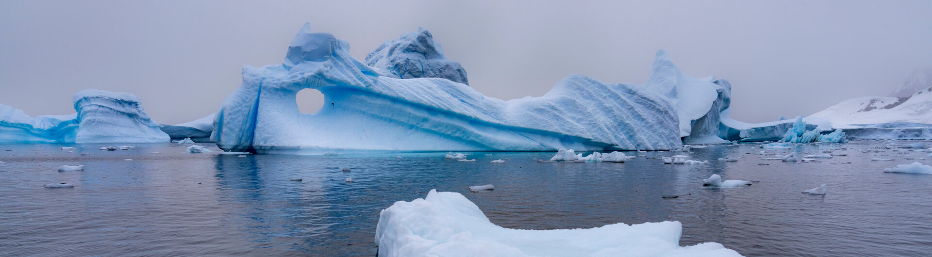 Ein kleinerer Eisberg treibt im Vordergrund im Meer vor einer großen Eisformation mit natürlichem Loch in der Antarktis.
