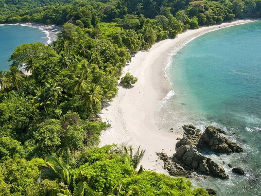 Blick von oben auf eine idyllische Bucht mit türkisfarbenem Wasser, weißem Sandstrand und üppigem Regenwald, Felsen im Vordergrund, weitere Bucht im Hintergrund in Manuel Antonio, Costa Rica