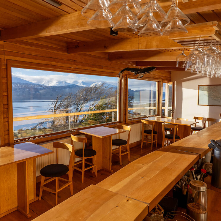 Holzrestaurant mit Bar und Tischen, große Panoramafenster mit Blick auf See und schneebedeckte Berge in Patagonien, Chile.