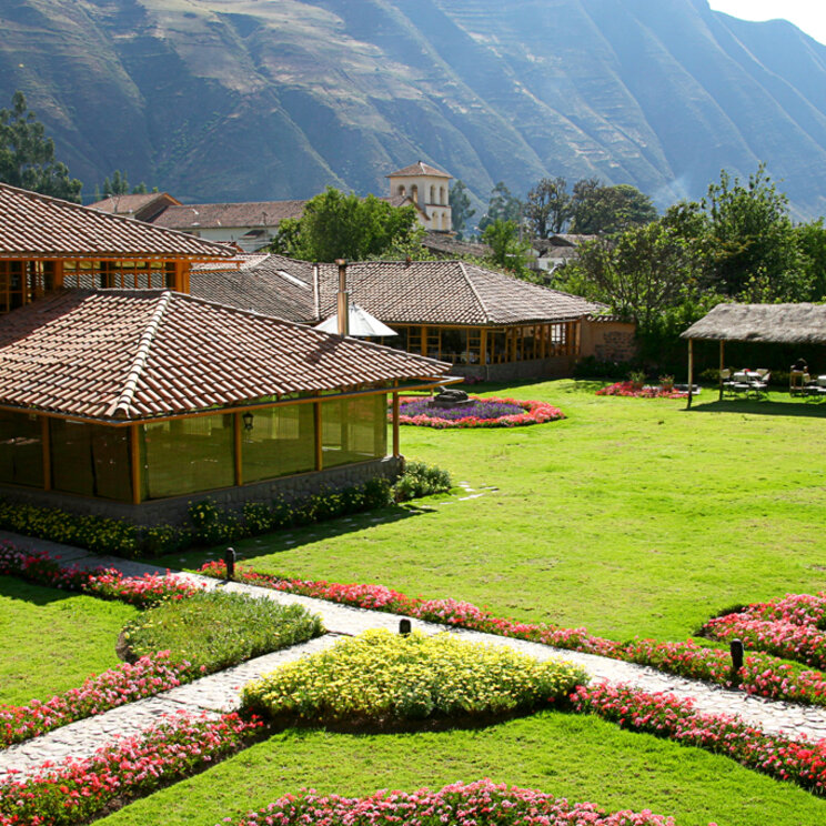 Gepflegte Gartenanlage mit Steinwegen, Blumenrabatten und strohgedecktem Pavillon vor Gebirgspanorama, Peru.
