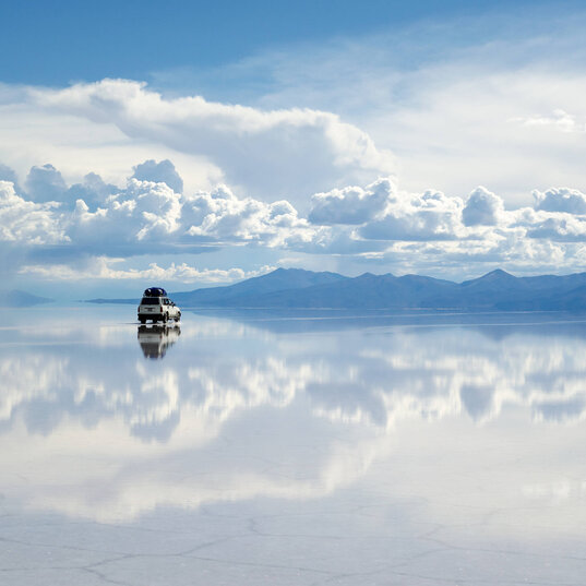 Auto steht auf der spiegelglatten Oberfläche des Salar de Uyuni während der Regenzeit. Der blaue Himmel und die Wolken spiegeln sich in der Salzwüste